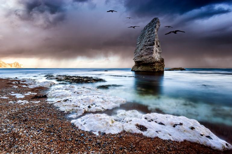 IDEE: Der Name Jurassic Coast für diesen Küstenabschnitt in Dorset rührt zwar von den Fossilienfunden her, aber der Fotograf schafft geschickt eine urzeitliche Atmosphäre. GESTALTUNG: Schöne Wiederholung des
Bogens der weißen Felsen vorn und der gekrümmten Felsnadel im Wasser. Die Vögel tragen die düstere Stimmung in das Foto. TECHNIK: Fünf Bilder mit Belichtungszeiten zwischen 1/200 und 59 Sekunden wurden
mit und ohne Graufilter angefertigt und in Photoshop manuell überblendet. Standort ist in der Nähe der Felsbrücke Durdle Door.