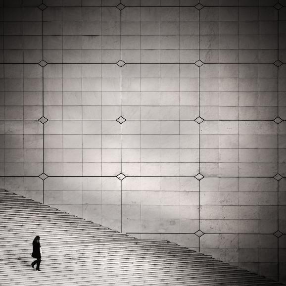 IDEE: Streetphotography als Architekturfoto: Die kleine Person scheint in der mächtigen Konstruktion des Grande Arche in Paris zu verschwinden und macht diese noch größer. GESTALTUNG: Exakt gesetzte Linien dominieren das Bild und stellen die Architektur in den Mittelpunkt. Sehr gut gefällt, dass das Umfeld aus dem Foto ausgeklammert wurde. TECHNIK: Zufallsfund im eigentlich sehr belebten Viertel La Défense, der Fotograf hatte nur ein paar Sekunden. Beschnitt und Vignette in der Bildbearbeitung durchgeführt. 