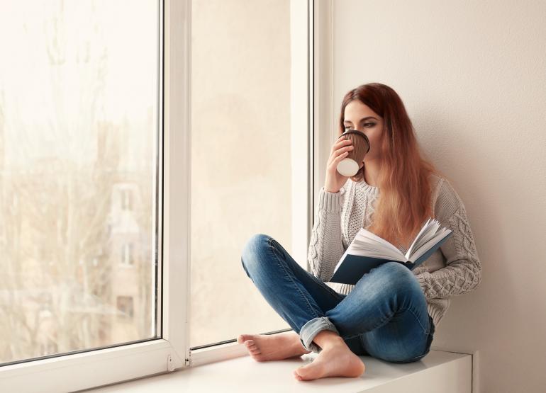 Frau trinkt Kaffee und liest ein Buch, während sie zu Hause auf der Fensterbank sitzt