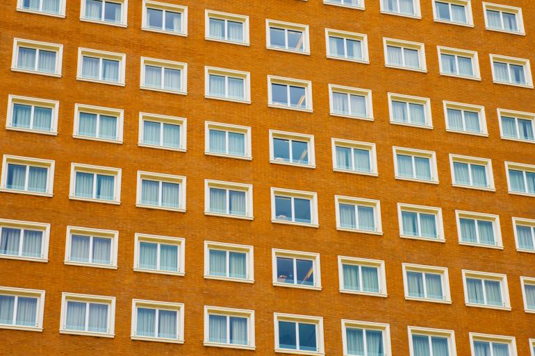 Die streng strukturierte Fassade eines Gebäudes wirkt durch Wiederholung und Symmetrie fast wie ein grafisches Muster. Nur wenige geöffnete Fenster durchbrechen die strenge Ordnung und geben der Architektur eine subtile Dynamik.