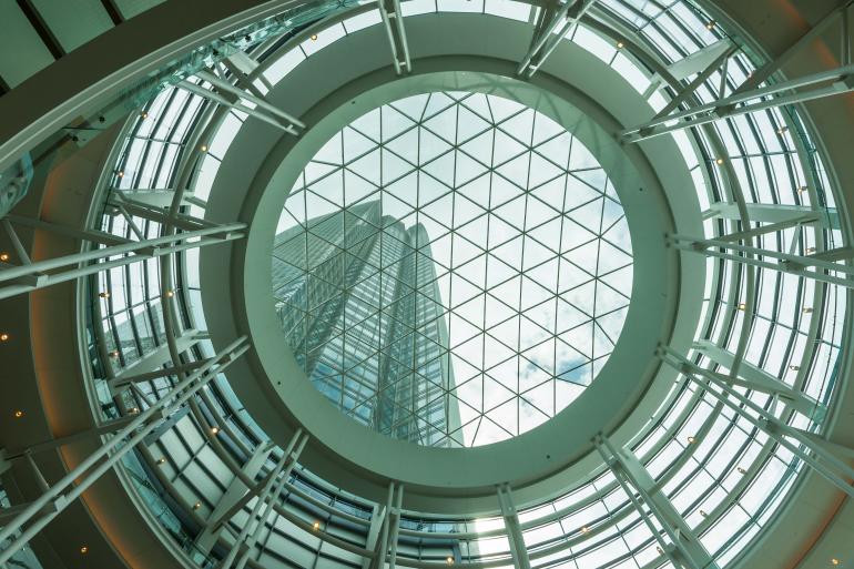 Ein architektonischer Blick nach oben: Die Glaskuppel mit ihrer geometrischen Struktur rahmt den Blick auf das darüberliegende Hochhaus und lässt Linien und Licht in perfekter Harmonie verschmelzen.